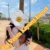 Person outdoors holding a book, a large white flower covers their face; orange diagonal watermark across the image.